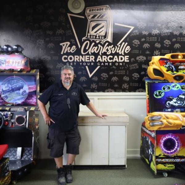 A man stands between arcade games in The Clarksville Corner Arcade, with a large arcade logo on the wall.