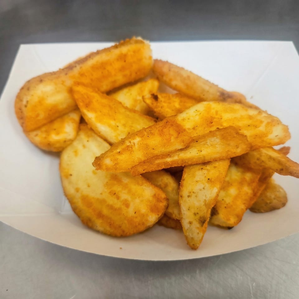 A white paper tray filled with golden-brown, seasoned potato wedges from The Clarksville Crust.