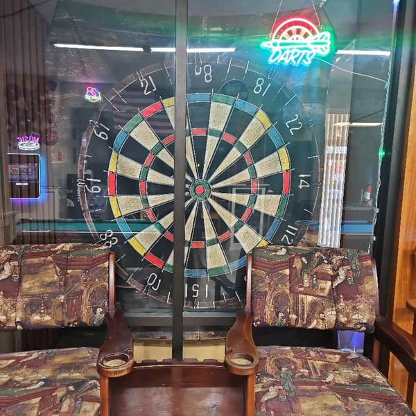 A dartboard with a neon 'DARTS' sign, seen through a window with patterned chairs in the foreground.