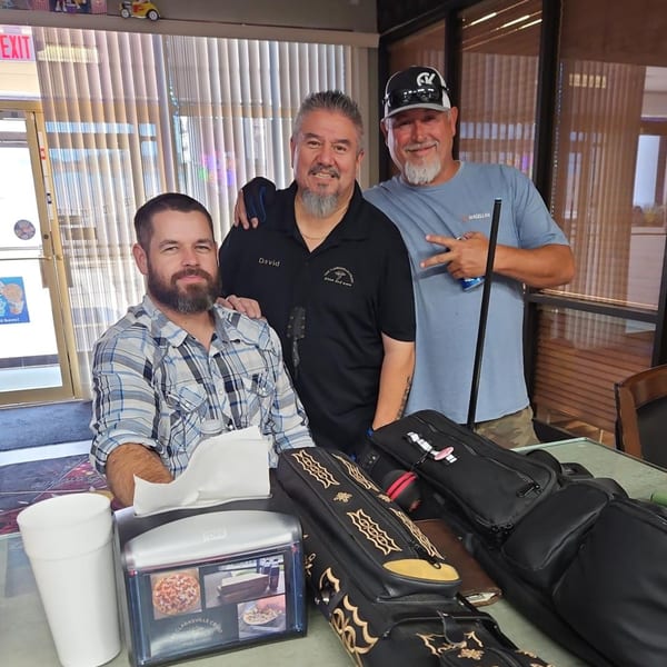 Three men, one wearing a "The Clarksville Crust" shirt, pose together indoors with pool cue cases.